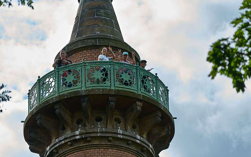 <p>Das von unten aufgenommene Foto zeigt oben auf der Galerie der Siegessäule stehende Frauen, die lachend nach unten winken.</p>
