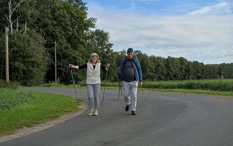 <p>Ein älteres Ehepaar wandert in sportlicher Kleidung und mit Wanderstöcken in der Hand über eine asphaltierte Straße, die von grünen Wiesen und Bäumen gesäumt ist.</p>