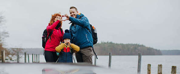 <p>Eine Familie aus Vater, Mutter und Kind steht am Seeufer, die Frau und der Mann bilden zusammen aus zwei Händen ein Herz.</p>