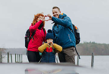 <p>Eine Familie aus Vater, Mutter und Kind steht am Seeufer, die Frau und der Mann bilden zusammen aus zwei Händen ein Herz.</p>