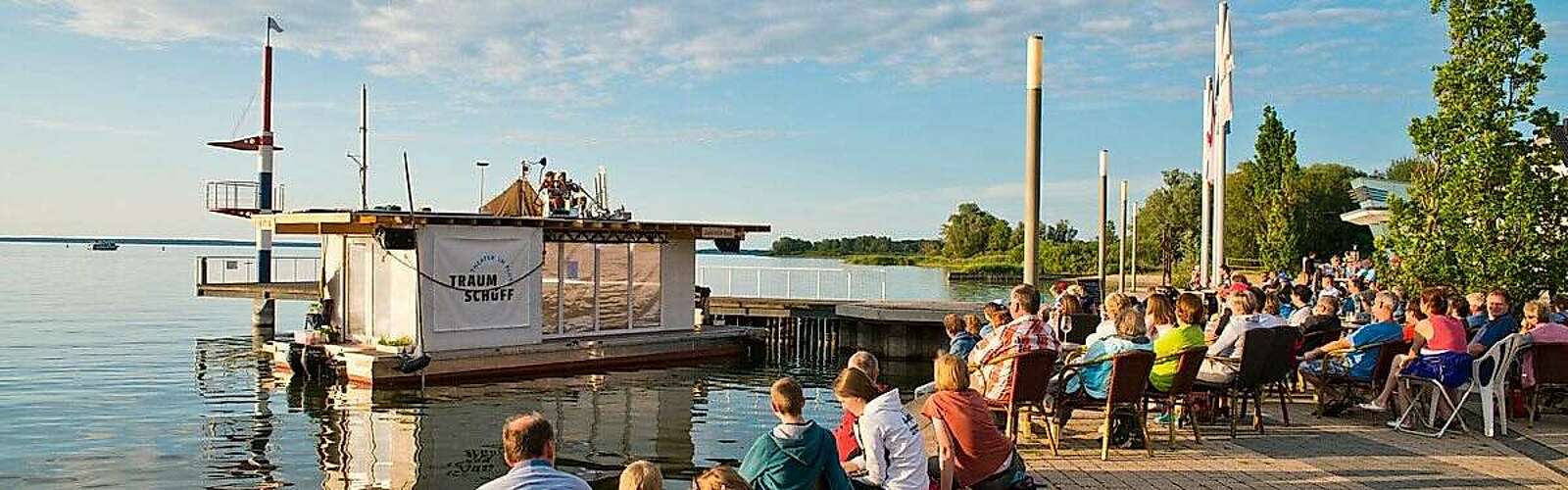 <p>Zwei Musiker sitzen auf dem Dach des Traumschüffes, einer mobilen Bühne auf dem Wasser. Zuschauer sitzen am Ufer.</p>