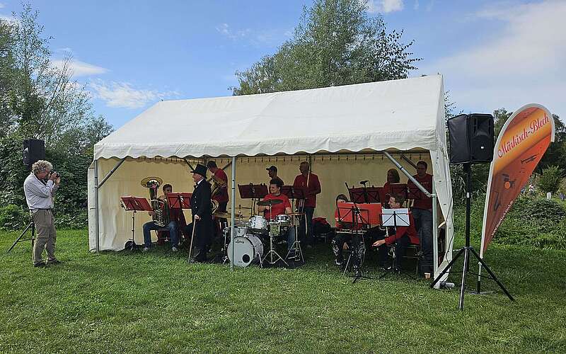 <p>Ein Mann fotografiert die unter einem Veranstaltungszelt sitzende, in rot gekleideten Mitglieder der Blaskapelle.</p>