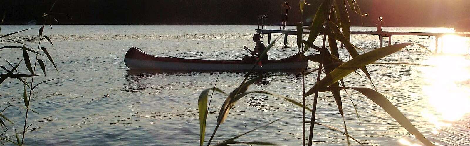 Eine Person fährt in der Abenddämmerung Kanu. Die untergehende Sonne spiegelt sich im Wasser.