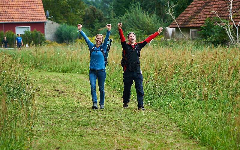 <p>Eine Frau und ein Mann wandern über einen ins grüne Feld gemähten Weg. Sie strecken freudig die Arme in die Luft und lachen.</p>