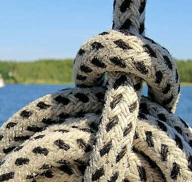 <p>Ein Seemannsknoten in einem weißen Seil mit schwarzen Punkten. Im Hintergrund sieht man einen weiten See mit waldigem Ufer und mehrere Boote.</p>