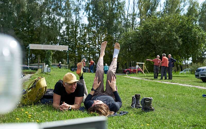 <p>Ein Mann und eine Frau liegen in sportlicher Kleidung auf einer Wiese, die Beine in die Luft, und ruhen sich aus.</p>
