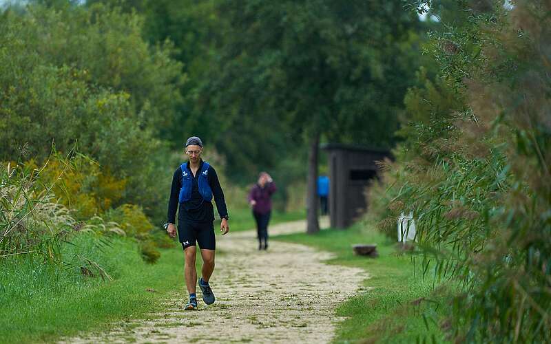 <p>Ein sportlich gekleideter junger Mann wandert auf einem leicht sandigen Weg entlang von Schilf und Bäumen.</p>